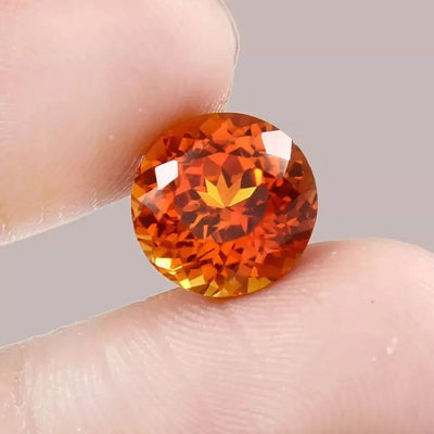 Round Orange Sapphire gemstone held between two fingers against a neutral background