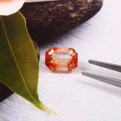 Bi-Color Unheated Padparadscha sapphire gemstone with tweezers and a leaf on a textured surface