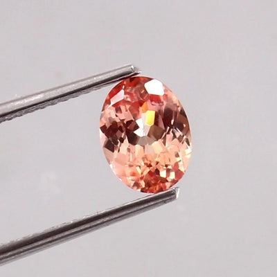Padparadscha Sapphire gemstone held by tweezers on a light gray background