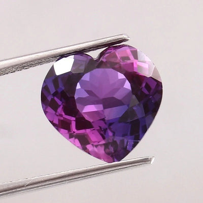 Heart-shaped Purple-Pink Sapphire gemstone held by tweezers on a light gray background