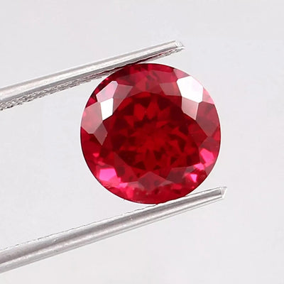 Round red Mozambique Ruby gemstone held between tweezers on a light gray background
