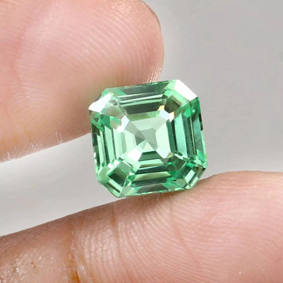 Green sapphire asscher cut gemstone held between two fingers against a neutral background